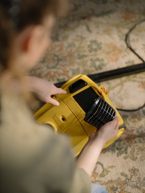 A person operating a yellow industrial carpet cleaning machine on a patterned area rug, which shows signs of dust and dirt. The machine has a black vent and control panel, and is connected to a power 