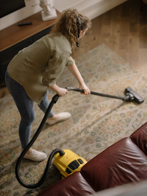 A person operating a yellow industrial carpet cleaning machine on a patterned area rug, which shows signs of dust and dirt. The machine has a black vent and control panel, and is connected to a power 